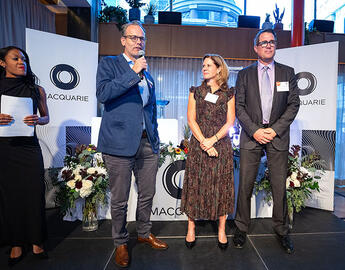 Aarnoud van Weelderen presenting from a stage at the The Macquarie Mix-Off event, with Lindsey McKay and Trevor Johnson beside him.