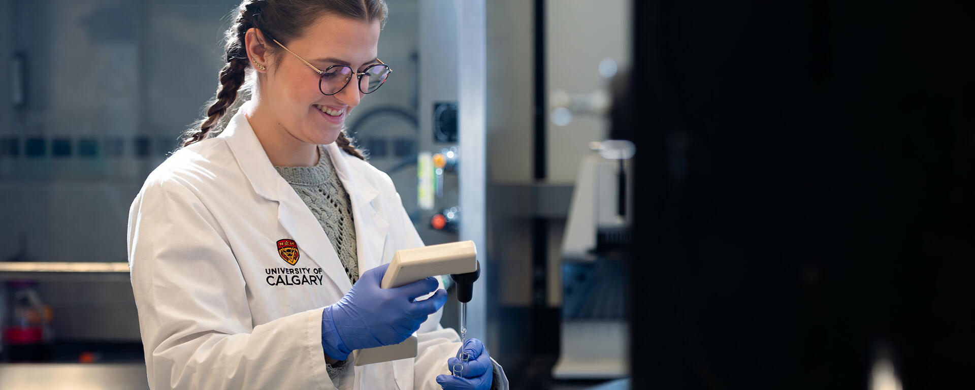 A young woman is working in a lab, wearing a University of Calgary lab coat.