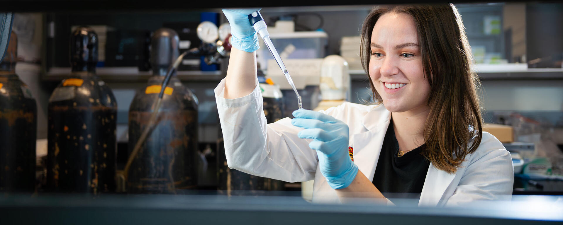 A young woman is working in a lab, wearing a University of Calgary lab coat.