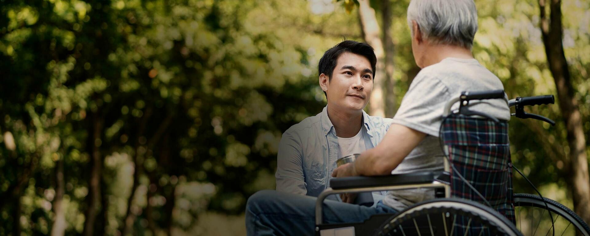 A young man looks at a senior man in a wheelchair while crouched beside him.