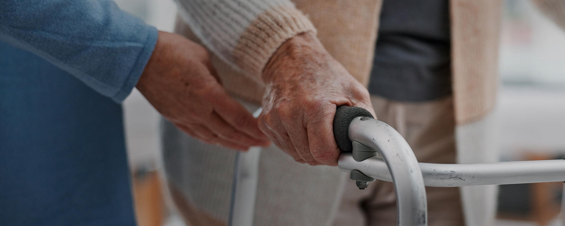 A caregiver's hand is placed close to a senior woman's hand on a walker.