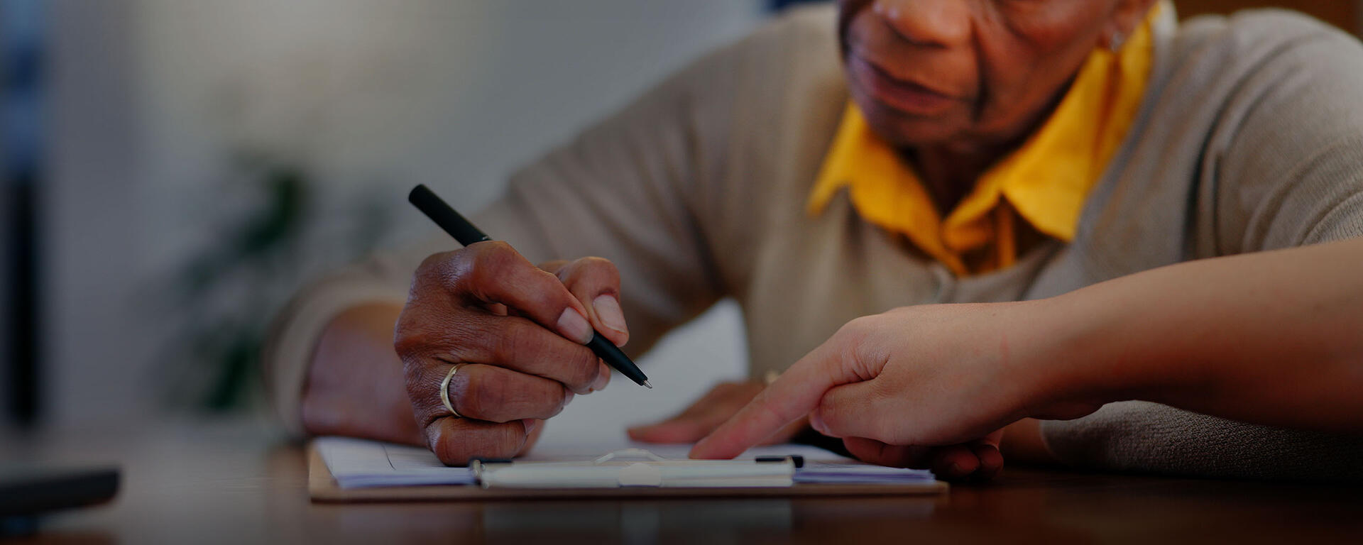 A senior woman signs documents, while a person points to the papers to help.