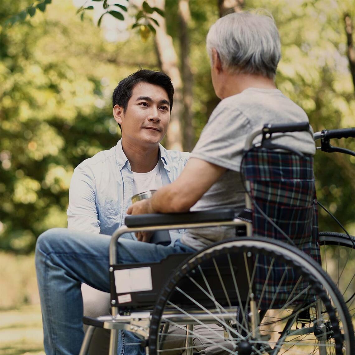 A young man looks at a senior man in a wheelchair while crouched beside him.