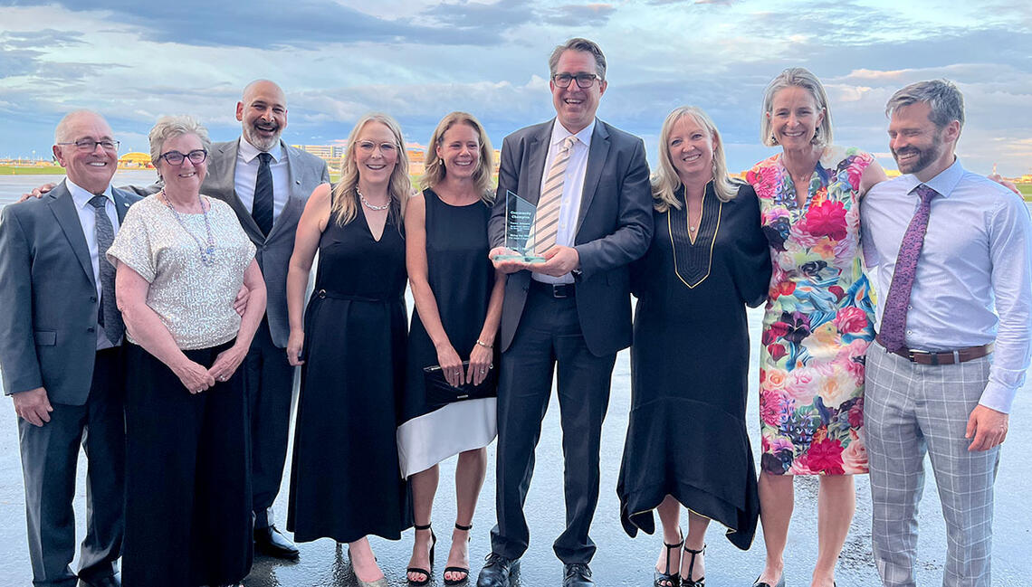 Trevor Johnson and Lindsey McKay stand beside each other, with a group of people on either side. Trevor is holding his Community Champion Award.