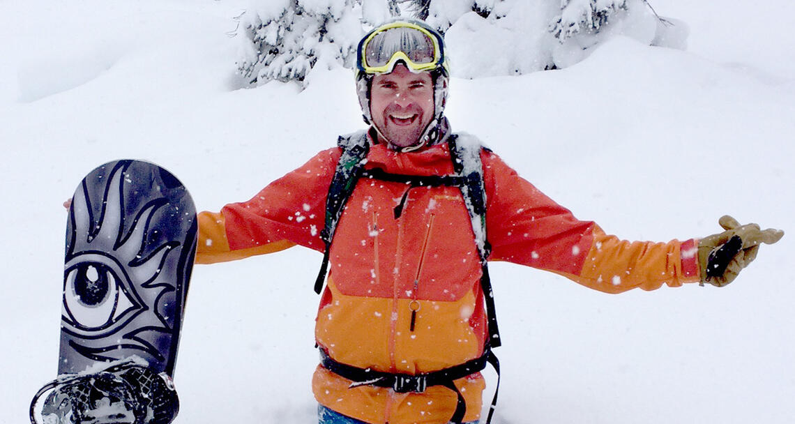 Mike McClay in the snow, smiling with his arms stretched out, holding a snowboard.