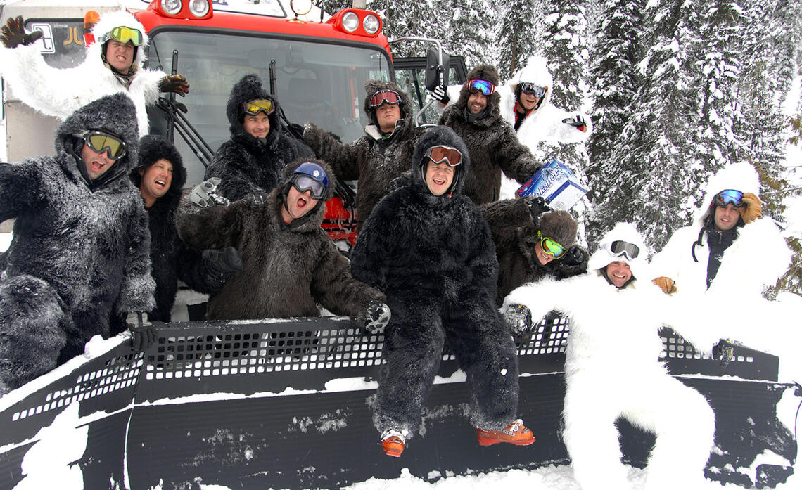 Trevor Johnson and Mike McClay cat skiing with a group of friends. Everyone is wearing black or white furry snowsuits and sitting on a snowcat.