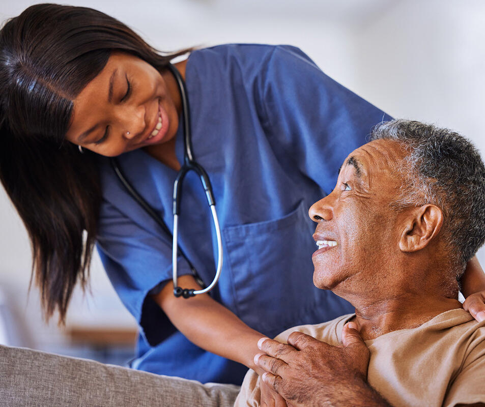 A nurse leans over with her hands placed on her patient's shoulders while they talk.