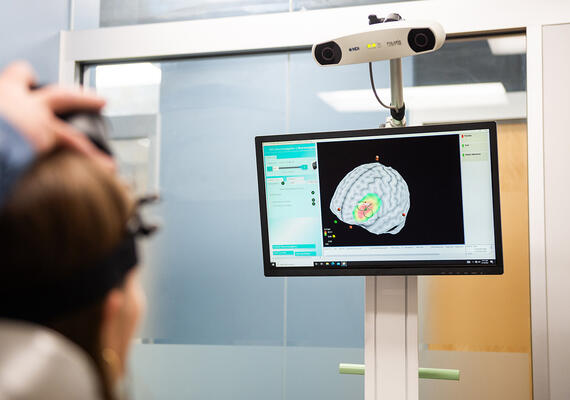 Dr. Alex McGirr demonstrates transcranial magnetic stimulation (TMS) on his graduate student, showing a depiction of her brain on a monitor.
