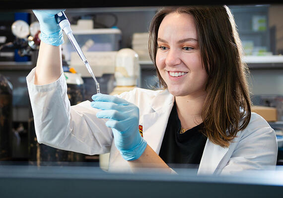 A young woman is working in a lab, wearing a University of Calgary lab coat.