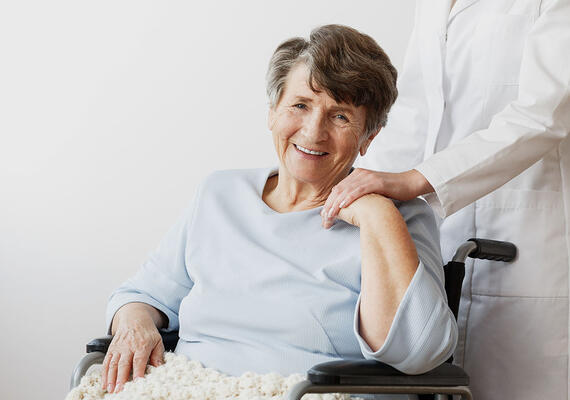 A senior woman smiles as a person in a lab coat puts their hand on her shoulder.