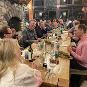 A large group of people are dining, with glasses of wine and appetizers placed on a long wooden table.
