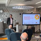 A group of people sit and listen to Trevor Johnston speaking, beside a screen that reads” Mental Health Initiative for Stress and Trauma (MIST).”