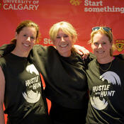 Three women with their arms around each other, stand in front of a University of Calgary backdrop.