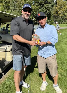 Two men on a golf course, posing side-by-side, holding a bag of Haribo Goldbears candy.