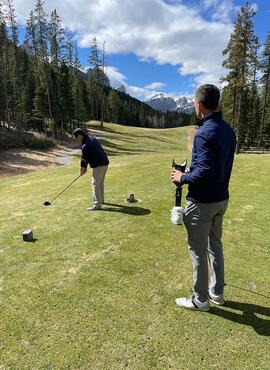 Two men are on a golf course, one is golfing and one is holding a prosthetic leg.