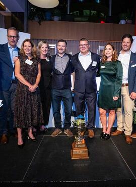 A group of people stand side-by-side, posing for the camera, with a trophy on the floor in front of them.