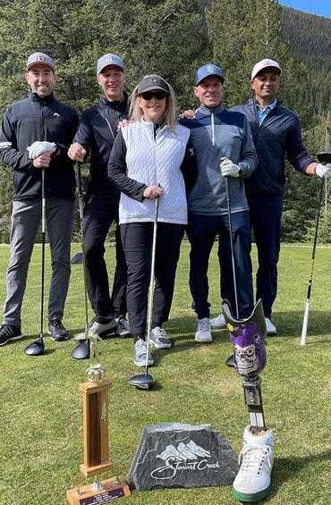 Five people posed on a golf course, holding golf clubs, in front of a trophy.