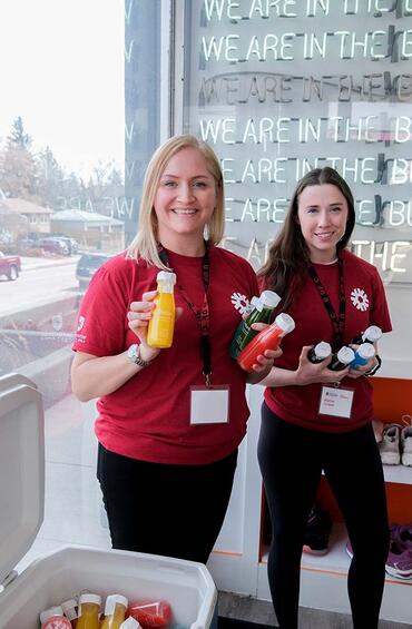 Two women are posing, wearing University of Calgary t-shirts and holding bottles of vibrant-coloured juices.