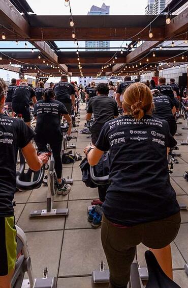 Looking at the back of a group of people on spin bikes, wearing Hustle with Hunt T-shirts.