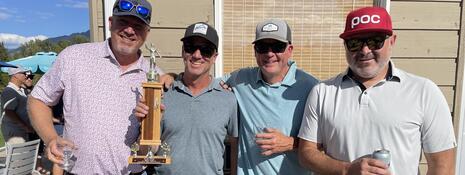 Four smiling men stand beside each other, posing for the camera, and one man is holding up a trophy.