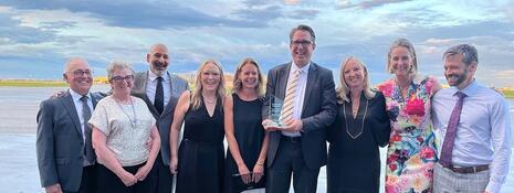 Nine people stand side-by-side outside, posing for the camera, with Trevor Johnston in the centre holding an award.