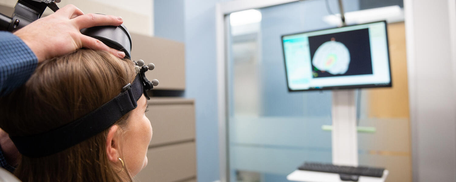 Dr. Alex McGirr demonstrates transcranial magnetic stimulation (TMS) on his graduate student, showing a depiction of her brain on a monitor.