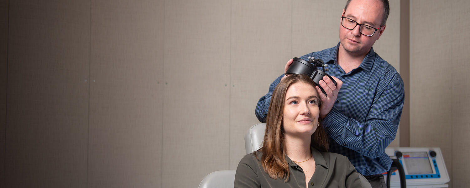 Dr. Alex McGirr demonstrates transcranial magnetic stimulation (TMS) on his graduate student, Jaeden Cole. McGirr is holding a small black device up against Jaeden's head.