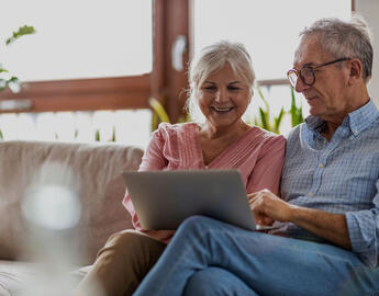 A senior couple is sitting on the couch looking at a laptop.