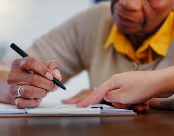 A senior woman signs documents, while a person points to the papers to help.